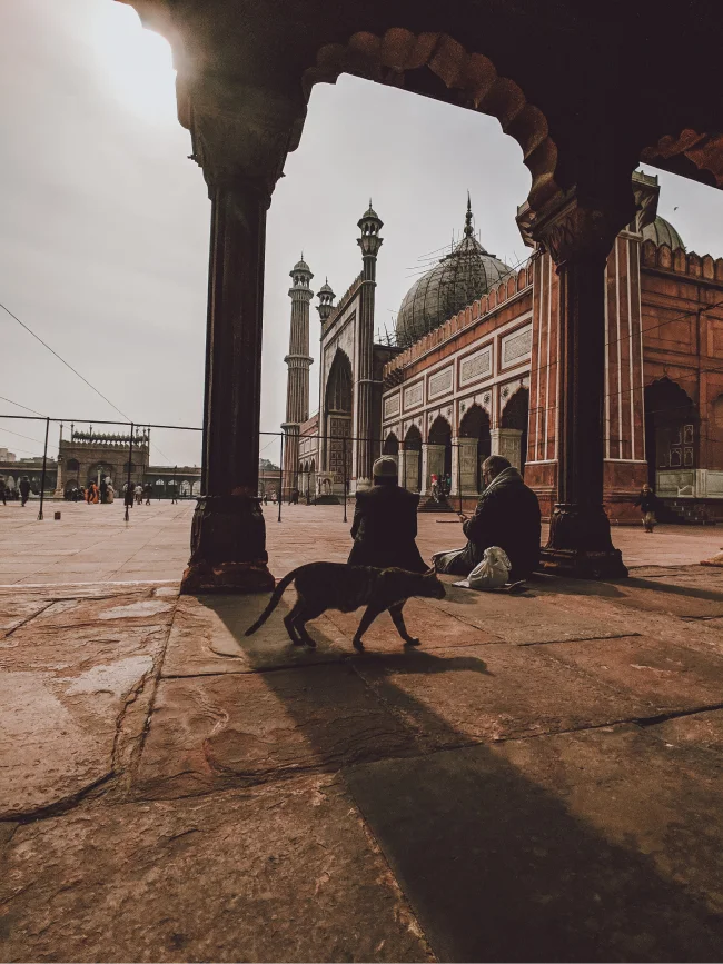 wo men seated in the courtyard of Jama Masjid in Delhi during sunset, with warm golden light illuminating the grand mosque's intricate architecture and serene ambiance