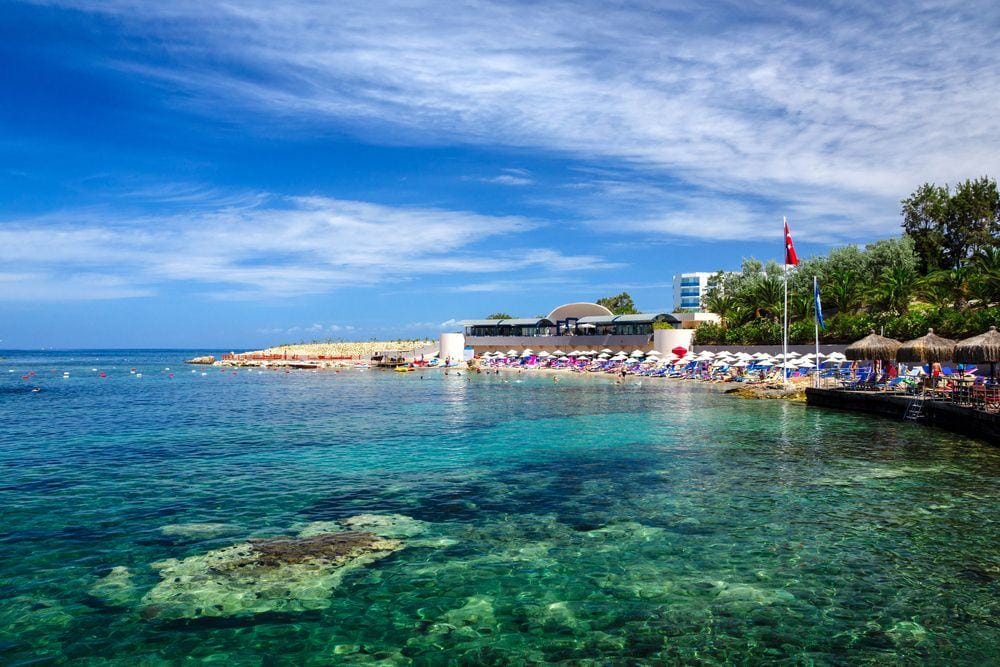 A view of Kusadasi beach with crystal-clear turquoise waters under a bright blue sky. The Turkish flag waves above the shoreline, colorful umbrellas line the soft sandy coast, and nearby buildings and lush greenery enhance the beauty of the scene