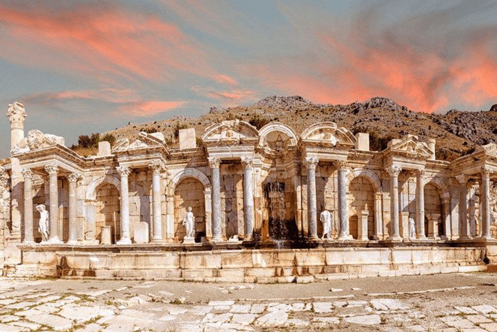 Ancient ruins of the Nymphaeum in Sagalassos, Turkey, with intricately carved arches and statues, set against a backdrop of rocky hills and a vibrant sunset sky